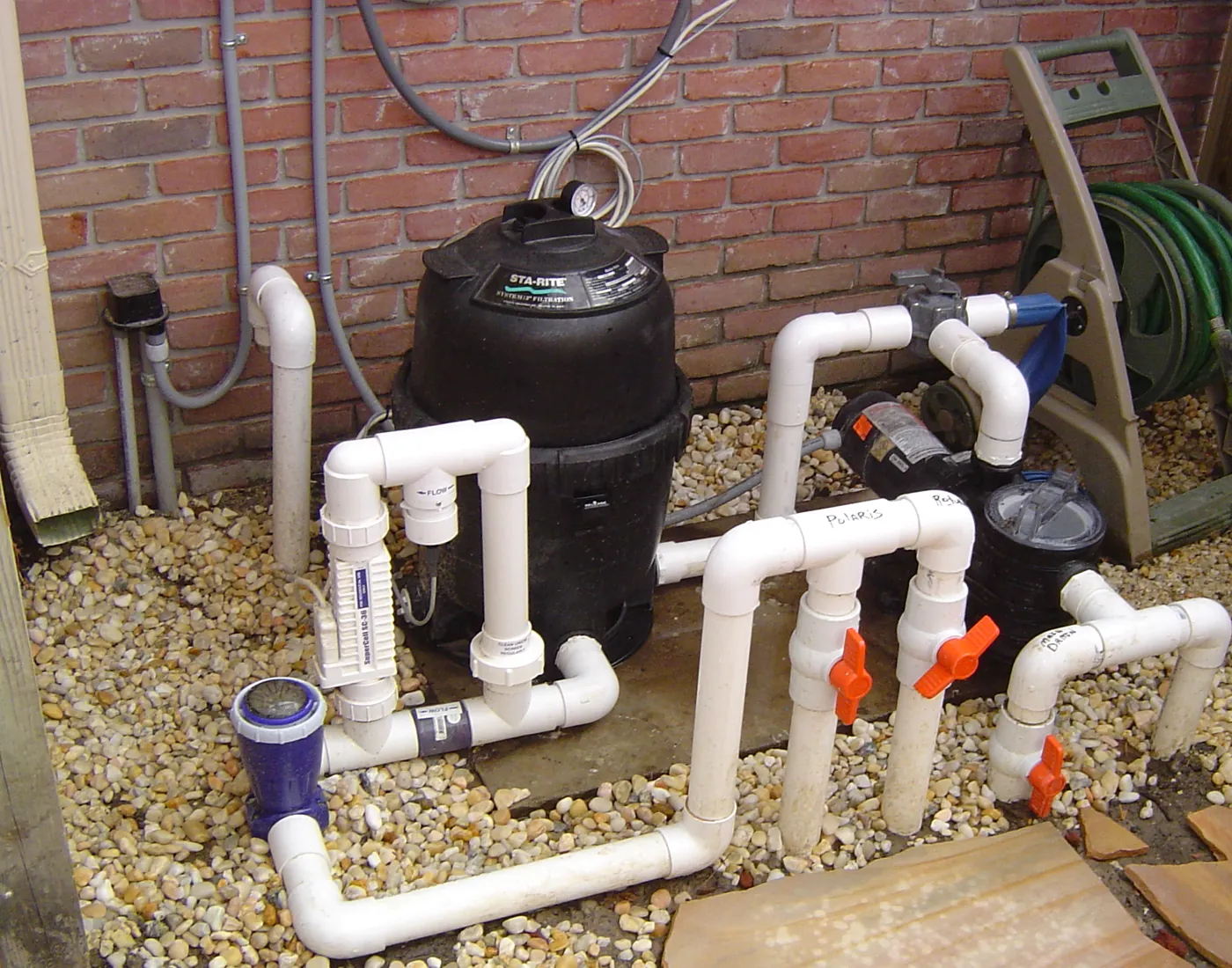 Residential irrigation system pump and PVC manifold assembly installed on a gravel bed beside a brick wall